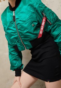 Groene bomberjack met een glanzende stof, zwarte geribbelde tailleband, zijzak met ritssluiting en een rode "Remove Before Flight" tag. Zwarte rok eronder.