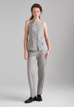 Femme aux cheveux bouclés portant un haut sans manches gris à fines rayures boutonné et un pantalon assorti avec des mocassins noirs, debout devant un fond uni.