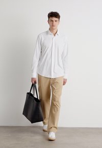 Hvit langermet skjorte, beige bukser og hvite joggesko. Svart tote-veske med to håndtak. Modell går på grått gulv mot en enkel bakgrunn.