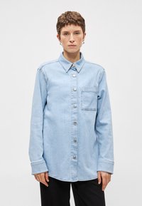 Ανοιχτό μπλε τζην πουκάμισο με κλασικό γιακά, μακριά μανίκια, μπροστινό κλείσιμο με κουμπιά και θηλάκι στο στήθος. Διαθέτει ασημί κουμπιά και ελάχιστες λεπτομέρειες.