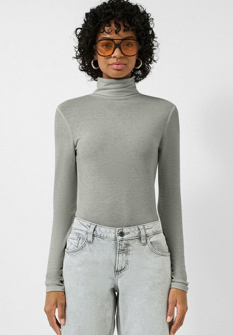 Femme aux cheveux bouclés portant de grandes lunettes teintées orange, des boucles d'oreilles argentées en forme d'anneau, un col roulé gris et un jean gris clair, debout devant un fond blanc.