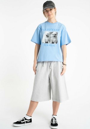 Młoda osoba w niebieskim T-shircie z nadrukiem puchatego psa, szarych szerokich szortach, czarnej czapce, czarnych trampkach z białymi sznurowadłami, stojąca na białym tle.