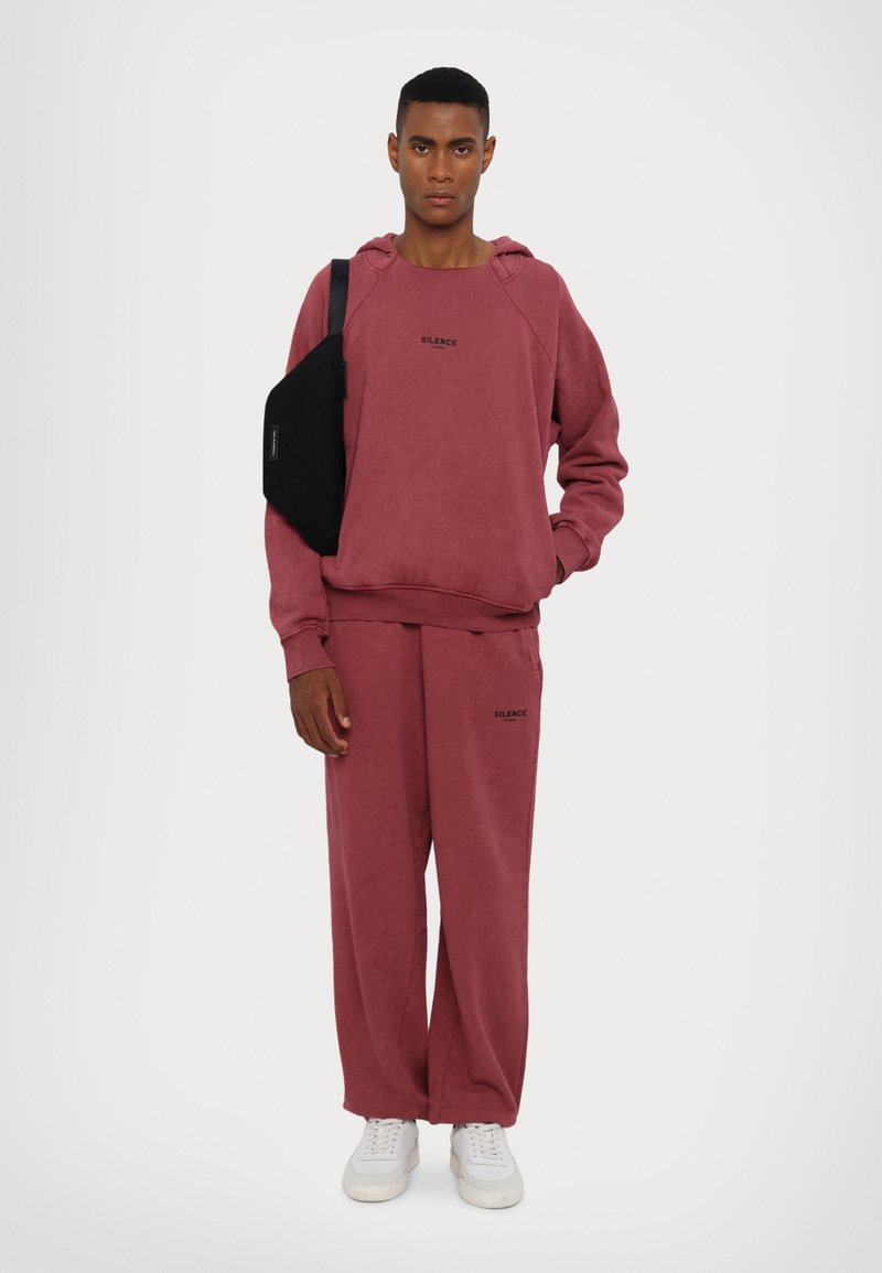 Sweatshirt com capuz cor bordeaux e calças de fato combinando, com um corte largo, apresentando o texto "SILENCE". Bolsa cruzada preta e ténis brancos.
