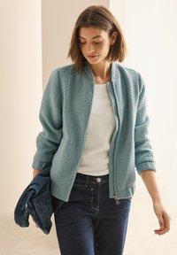 Giacca texturizzata blu chiaro con zip frontale, polsini a costine e scollo rotondo, abbinata a una maglietta bianca e jeans in denim scuro.