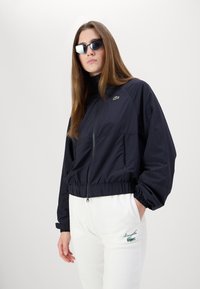 Tamsiai mėlynės zip-up vėjo striukė su elastiniais džemperių rankogaliais ir apačia, papuošta mažu žaliu logotipu. Derinama su baltomis treningų kelnėmis su žalia siuvinėjimu.