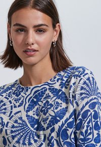 Sinivalkoinen kuvioitu pusero, jossa on kukka- ja geometrisia kuvioita, valmistettu kevyestä kankaasta, pyöreällä kaula-aukolla ja lyhyillä hihoilla.