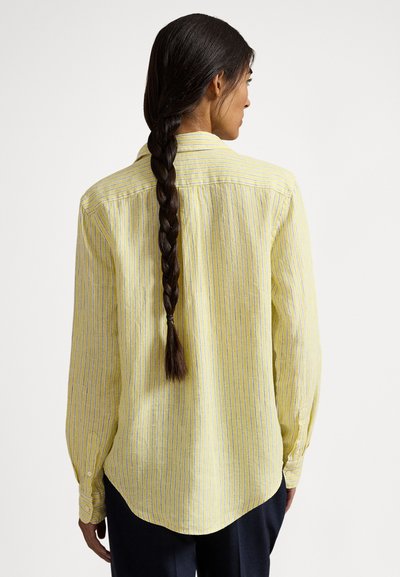 Sieviete ar muguru skatoties, gara tumša pinumā sasprausta matu frizūra, valkājot dzeltenu svītrainu garroku kreklu un tumšas bikses uz vienkārša fona.