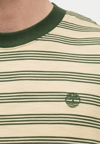 Κοντινή λήψη ενός μπλουζιού με ρίγες σε μπεζ και πράσινο χρώμα, με πράσινο ριμπ ύφασμα στον γιακά και ένα μικρό κεντημένο λογότυπο δέντρου στο στήθος.