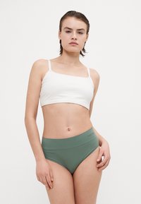 Biely crop top s štvorcovým výstrihom a zelené nohavičky s vysokým pásom. Tkanina vyzerá hladko s minimálnymi švami. Diskrétny branding na páse.