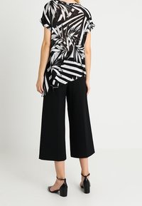 Czarna i biała tunika z wzorem palmowych liści, krótkie rękawy, asymetryczny dół, zestawiona z czarnymi spodniami w szerokim kroju i sandałami na wysokim obcasie.