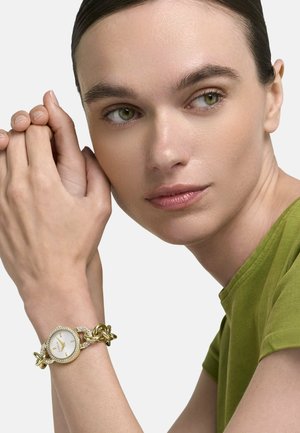 Femeie cu părul dat pe spate, purtând un ceas-brățară cu zale aurii și o cămașă verde măsliniu, cu mâinile încleștate odihnindu-se aproape de față.