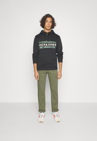 Black hoodie with green and white logo band, drawstring hood, cotton material. Olive green pants with rolled cuffs. White sneakers with red accents.