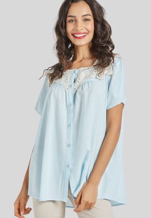 Mujer con cabello oscuro y rizado que lleva una blusa azul claro suelta con botones y detalles de encaje, sonriendo frente a un fondo liso.