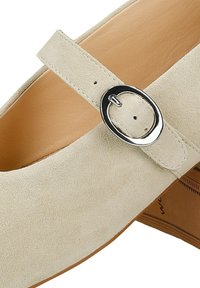 Chaussure Mary Jane en daim beige avec une bride à boucle ronde argentée, une doublure intérieure en cuir lisse marron clair et une semelle marron clair avec un petit talon.