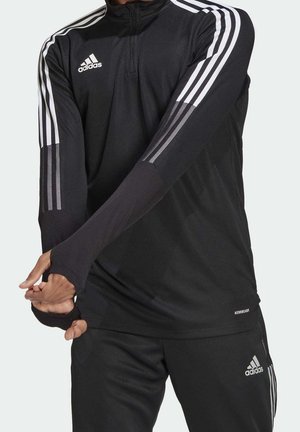 Personne portant un survêtement noir Adidas avec des bandes blanches sur les bras et les jambes, tirant une manche longue sur la main.