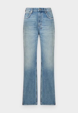 Nainen, jolla musta toppi, vaaleansiniset farkut sivulla niiteillä, mustat sandaalit ja isot mustat aurinkolasit, seisoo käsi taskussa.