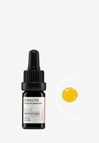 Sérum Odacité Cumin Noir-Cajeput dans un flacon en verre noir avec un compte-gouttes, dotée d'un capuchon strié et d'une étiquette rose. Goutte de sérum jaune à côté.