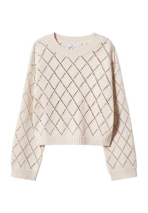 Person med langt sort hår iført beige strikket trøje med diamantmønster og sorte bukser, stående med front mod en ensfarvet lysegrå væg.