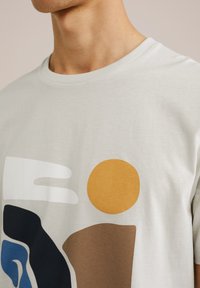 Světle šedé tričko s abstraktními tvary v námořnické modři, hnědé, bílé a oranžové barvě. Měkký bavlněný materiál s kulatým výstřihem.