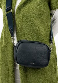 Borsa a tracolla in pelle nera con superficie texture, hardware argentato e tracolla regolabile, indossata sopra un cappotto verde texturizzato.