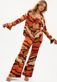Femme portant un chemisier à motifs abstraits orange vif, marron et beige, un haut court et un pantalon évasé, posant les bras tendus.