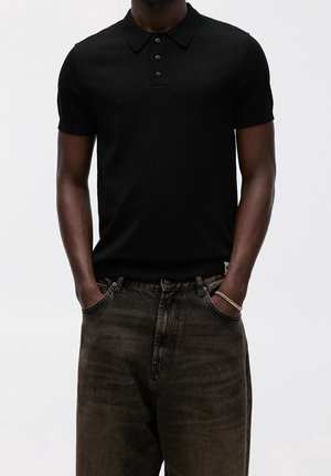 Homme portant un polo noir à manches courtes rentré dans un jean noir délavé, mains dans les poches et un bracelet argenté au poignet gauche.