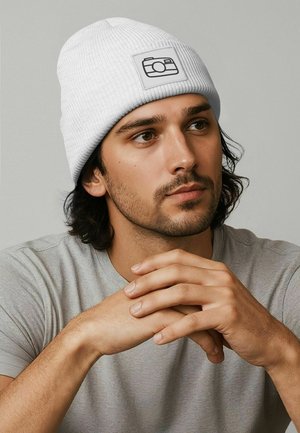 White ribbed beanie with a rectangular patch featuring a camera graphic, worn with a gray T-shirt, hands clasped in a neutral pose.