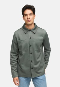 Grøn jakke med knaplukning og spredt krave, to brystlommer og sorte knapper; lavet af struktureret stof, sat sammen med blå jeans.