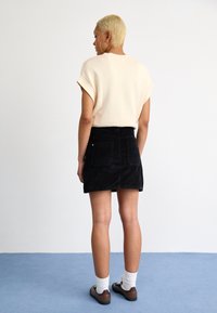 Pull court beige clair à manches courtes et mini jupe en velours côtelé noir. Le modèle porte des chaussettes blanches et des chaussures brunes, se tenant contre un fond uni.