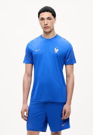Jeune homme debout portant le maillot bleu de football de la France avec le logo Nike et l'emblème de la FFF sur fond blanc uni.