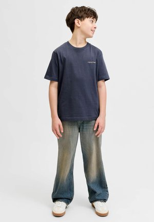 Garçon portant un t-shirt bleu marine, un jean ample délavé et des baskets blanches, regardant vers sa droite sur un fond uni.