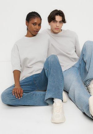 To unge voksne sidder på gulvet side om side iført lysegrå oversized toppe, blå jeans og hvide sneakers, mod en hvid baggrund.