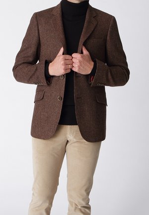 Man adjusting brown textured blazer worn over black turtleneck and beige pants against plain white background.