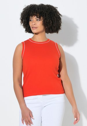 Femme aux cheveux bouclés portant un haut rouge sans manches avec bordure blanche et un pantalon blanc, debout devant un fond blanc.