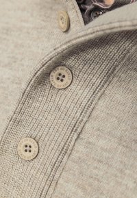 Beige gestrickter Pullover mit strukturiertem Finish, ausgestattet mit drei braunen Knöpfen, die mit "STATE OF ART" geprägt sind, und einem Kragen-Detail.