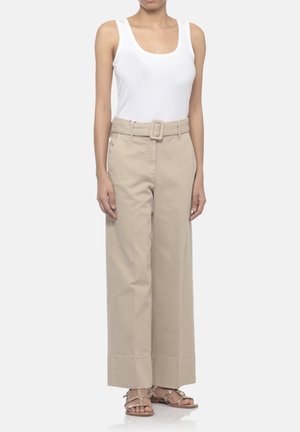 Donna che indossa un top bianco senza maniche, pantaloni beige a gamba larga e vita alta con cintura abbinata e sandali beige con borchie.