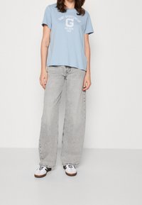 Lichtblauw katoenen T-shirt met korte mouwen en witte grafische tekst. Gecombineerd met brede, vervaagde grijze jeans en witte sneakers met zwarte accenten.