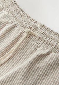 Gros plan sur une ceinture élastique beige et blanche à rayures avec un cordon blanc noué sur un short en tissu léger.