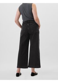Jean noir à jambes larges en denim, avec une taille haute, des poches arrière et un ourlet net. Porté avec un haut gris sans manches.