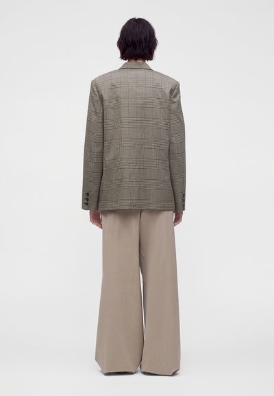 Personne vue de dos portant un blazer marron à carreaux et un pantalon large beige, debout devant un fond clair uni.