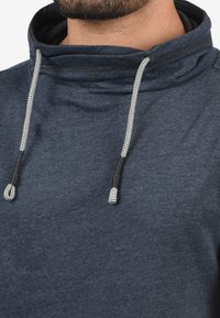 Marineblaues Hoodie mit hohem Kragen, strukturiertem Stoff und grauen Kordeln mit schwarzen Spitzen. Bietet ein lässiges, entspanntes Design.