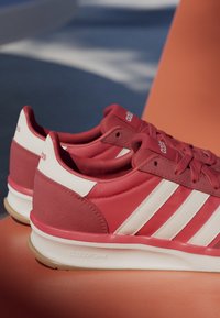 Paire de baskets Adidas rouges et blanches avec des bandes blanches et des semelles Cloudfoam reposant sur une surface orange.
