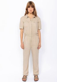 Combinaison en lin beige à manches courtes, fermeture boutonnée sur le devant, poches poitrine et jambes fuselées. Dispose d'une taille ceinturée et d'une coupe décontractée.