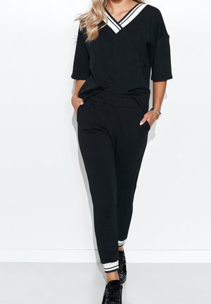 Vrouw draagt een zwart V-hals top met witte strepen en bijpassende zwarte joggingbroek met witte gestreepte manchetten, staand met handen in de zakken.