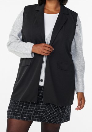 Mujer con blazer negro sin mangas sobre cárdigan gris claro y camiseta blanca, combinados con falda de cuadros en blanco y negro.