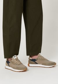 Baskets en suède beige et mesh avec semelles blanches, accents bleus et détails gris. Lacets et logo sur la languette visibles, portées avec un pantalon vert foncé.