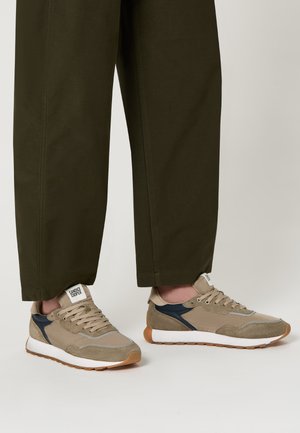 Sneakers in suede e mesh beige con suole bianche, dettagli blu e grigi. Lacci e logo sulla linguetta visibili, indossati con pantaloni verde scuro.