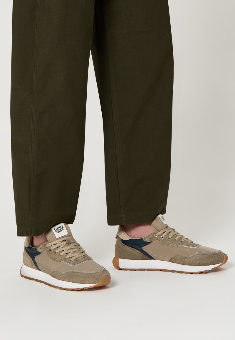 Baskets en suède beige et mesh avec semelles blanches, accents bleus et détails gris. Lacets et logo sur la languette visibles, portées avec un pantalon vert foncé.