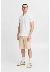 Valkoinen polo-paita, jossa on kaulus ja vaaleanbeige reunus, yhdistettynä vaaleanbeigeihin shortseihin. Kengissä on valkoisia ja beigejä yksityiskohtia.