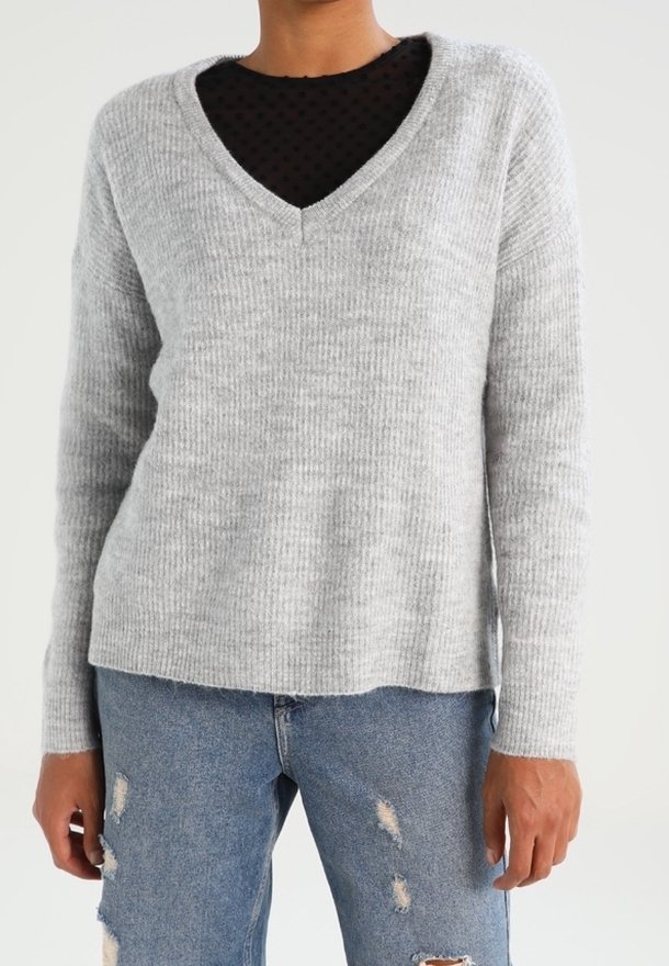 Maglione grigio a coste con scollatura a V, abbinato a una maglietta nera a pois sotto, indossato con jeans blu strappati.